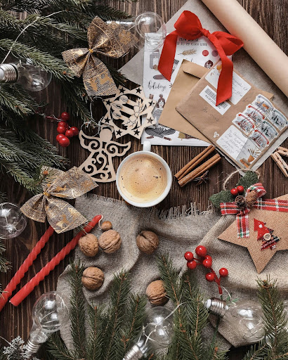 A cozy holiday flat lay featuring evergreen branches, gold bows, red candles, wooden ornaments, envelopes with festive stamps, cinnamon sticks, walnuts, and a cup of coffee arranged on a rustic wooden table.