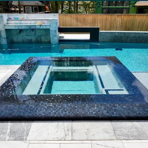 Close-up of a modern pool with a glass-tiled hot tub, marble accents, a waterfall feature, and a wooden privacy fence in the background.