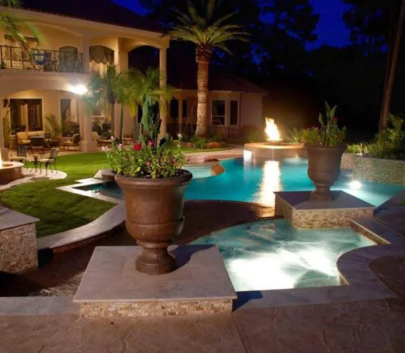 Night view of a luxury backyard with a swimming pool, fire feature, tropical plants, and a villa surrounded by elegant landscaping.