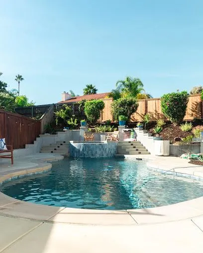 Daytime view of a backyard pool with a waterfall feature, terraced landscaping, potted plants, and a wooden privacy fence under a clear blue sky.