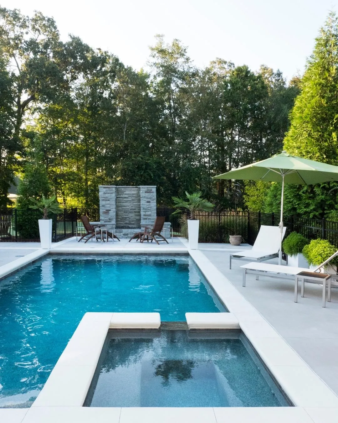 Modern rectangular pool with raised spa and stone water feature, surrounded by greenery and lounge chairs.
