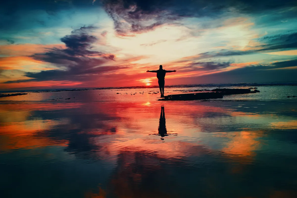 Silhouette of a person standing on a wet beach shore with a colorful sunset in the background