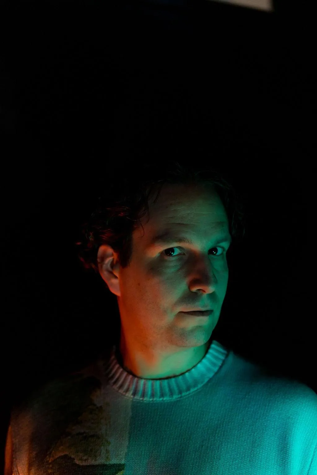 GrayBeat with arched eyebrows looks to camera with a side glance, a green glow on one side of the face and a red glow on the other. no sunglasses.