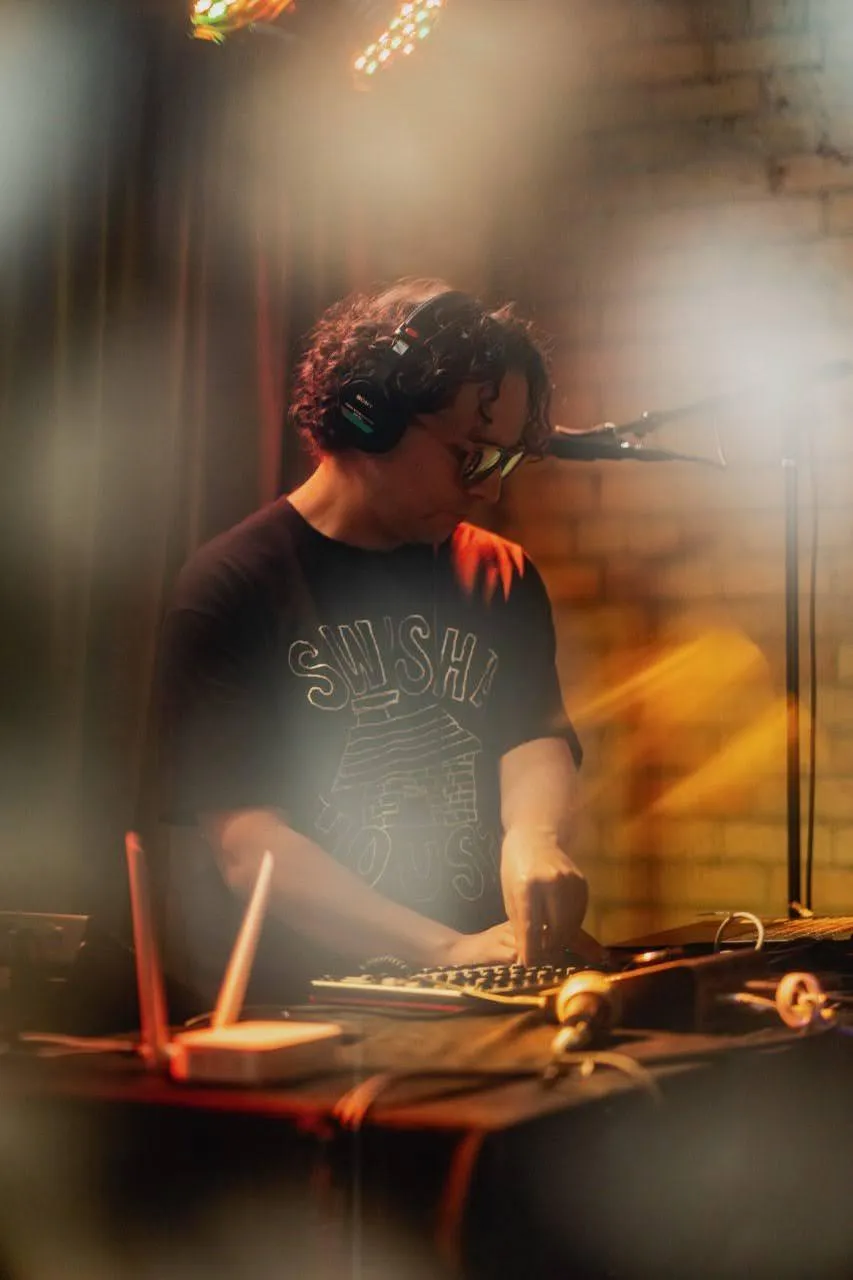 GrayBeat performs live at Underground Music Cafe, headphones & sunglasses on. adjusting sound and midi equipment on the table with a microphone to his side. a brick wall flanks graybeat, and a dreamy blur envolopes the whole scene.
