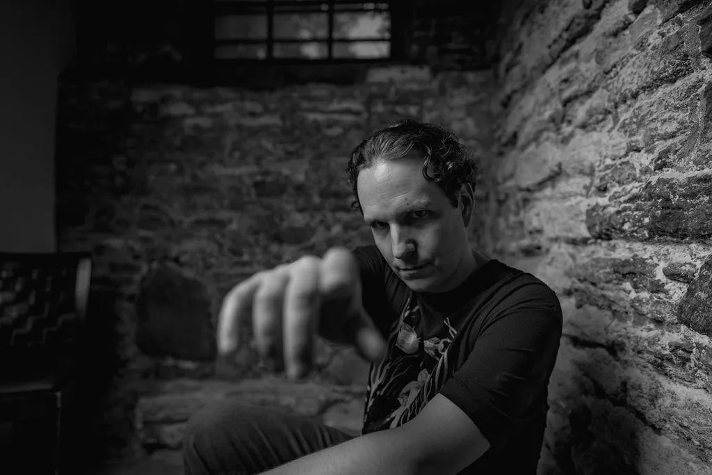 Robin extends his hand almost to the camera lens. sitting down, he turns to the camera and smiles. same brick studio, this time sitting position on a bench lining the wall, a relaxed manner. a knowing gaze.