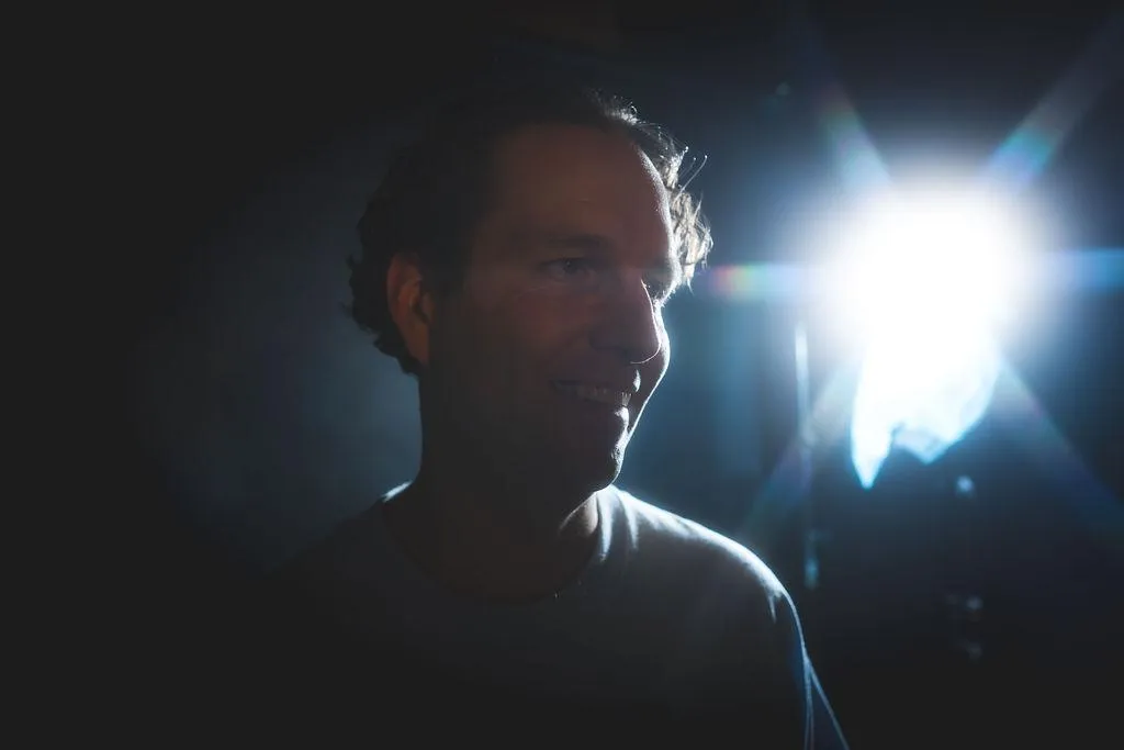 Robin in a full smile/laugh looking away from the camera, facing ahead. intense spotlight in the background shines on his cheek, with heavy shaddows and darkness otherwise. revealing a stark profile view of the artist.
