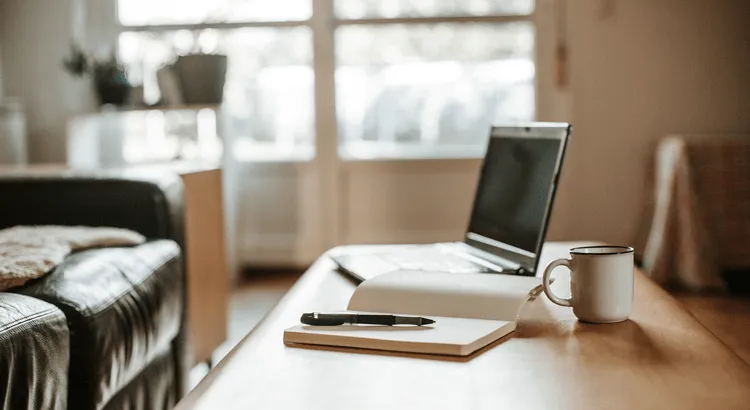 A laptop, book, pen and a cup of coffee on a desk in a well lighted room.