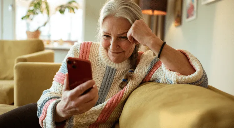 Woman sitting on a light brown couch inside her home happily looking at her mobile phone.