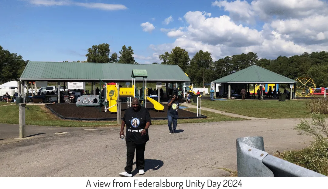 Another view of the Unity Day Celebration shows a man in an "I'M WITH HER" shirt walking across the street in front of a playground and a busy pavilion.