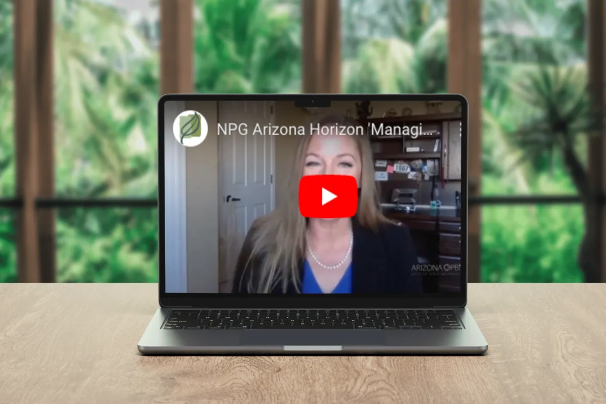 A laptop on a wooden desk displaying a YouTube video thumbnail from NPG Arizona Horizon. The video shows a woman with long blonde hair wearing a blue top and pearl necklace, speaking in what appears to be a home office. The laptop is set against a background of lush green foliage visible through large windows.