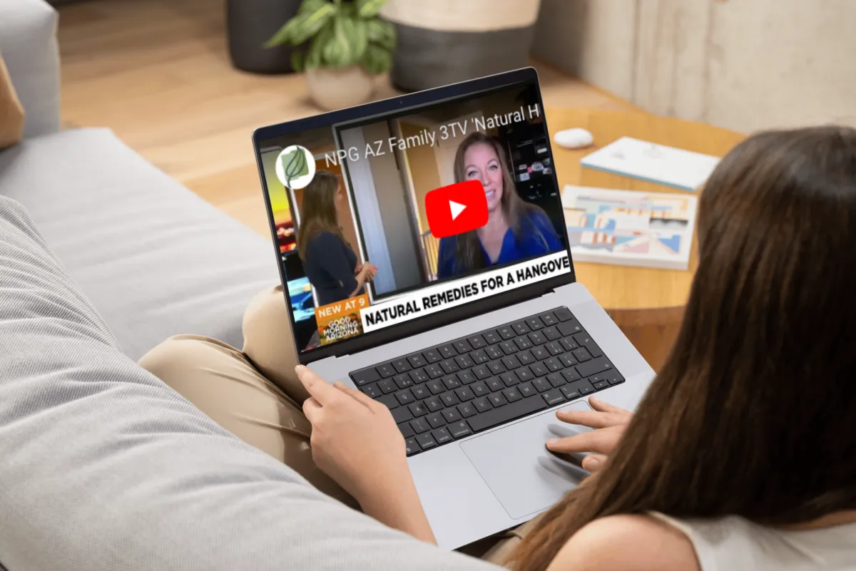 A person sitting on a couch, holding a laptop that displays a YouTube video thumbnail from NPG AZ Family 3TV about natural remedies for a hangover. The video shows two women, one in a blue top, discussing the topic. The laptop screen also features the NPG logo and a "Natural Remedies for a Hangover" caption.