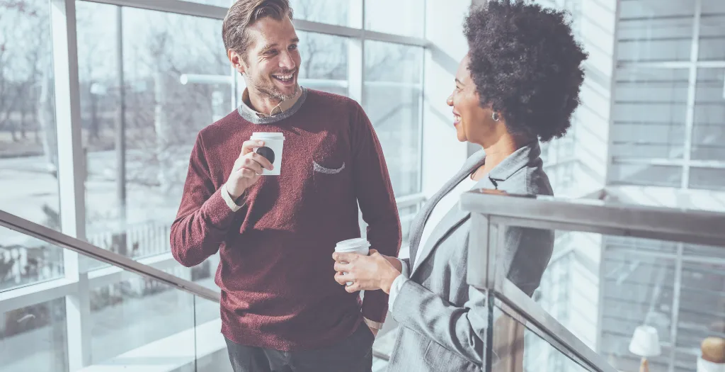 Two professionals smiling and chatting over coffee on a staircase in a sunlit office.