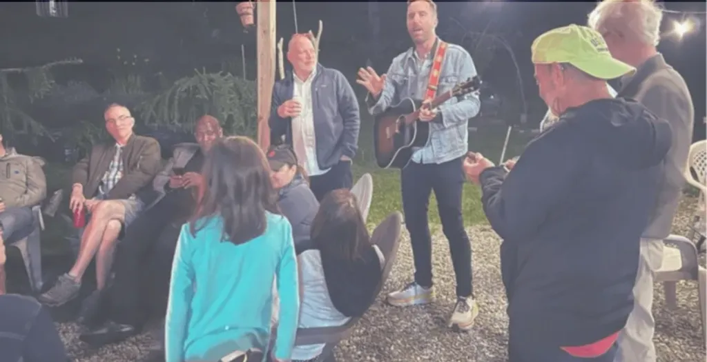Diverse group gathered outdoors at night, attentively listening to a man playing guitar and speaking.