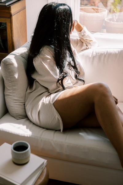 Woman sitting on a sofa in soft natural light, gazing out a window with a thoughtful expression, conveying quiet reflection and fatigue