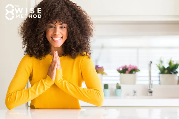 Woman in bright yellow jumper looking to camera smiling doing pray pose in her kitchen