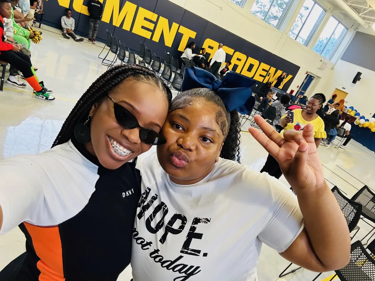 Koach Keisha posing with a young adult at what appears to be a school or community event. She is smiling warmly, dressed in a stylish and colorful outfit, while standing beside the young person, who is also smiling. The background suggests an indoor setting, possibly a hallway or event space, indicating a mentorship or coaching engagement.