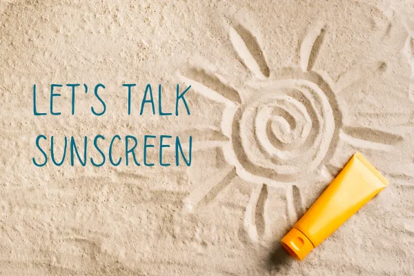 Sun drawn in the sand with a bottle of sunscreen and text title Let's Talk Sunscreen