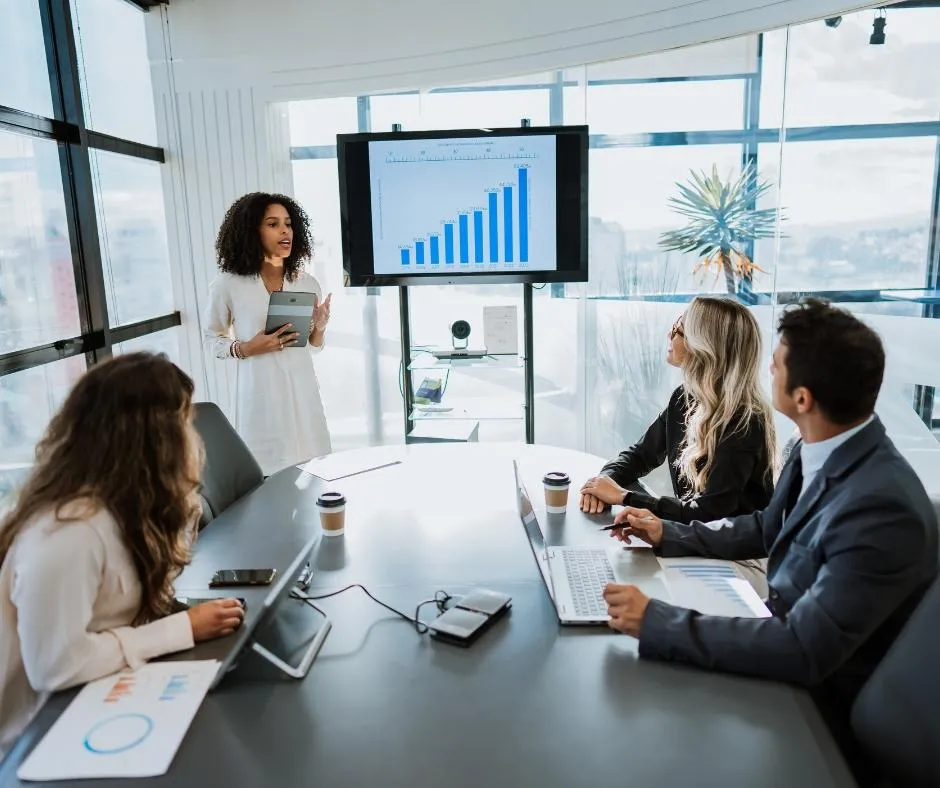 Woman presenting a rising bar chart to colleagues in a bright, glass walled conference room, holding a tablet