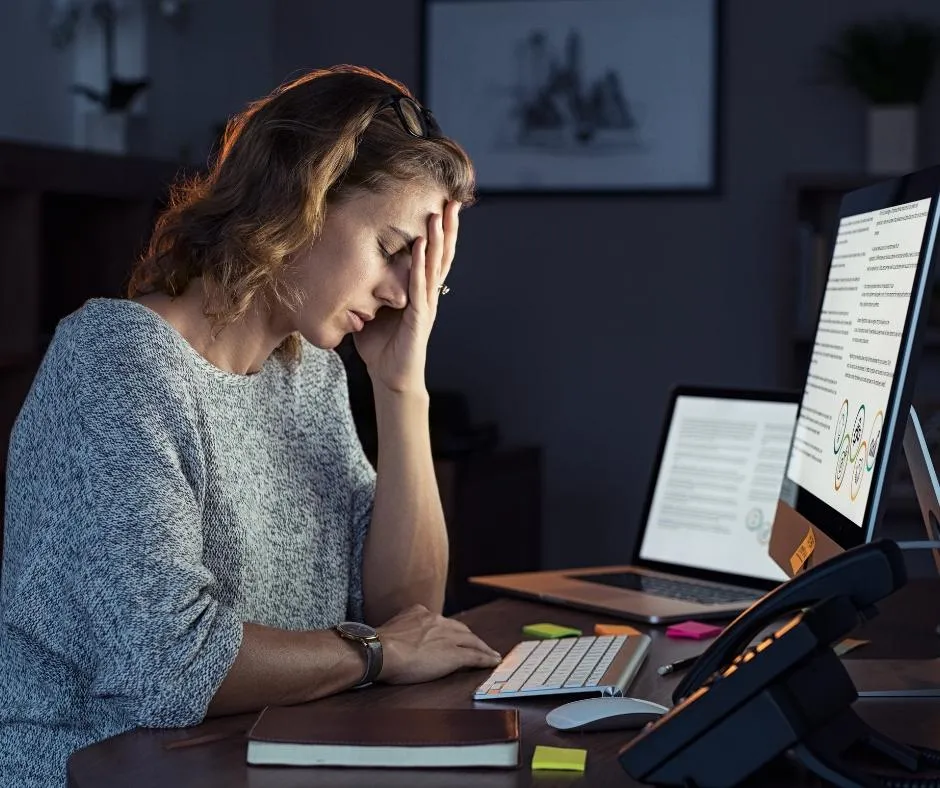 Stressed woman sitting at her desk with multiple computer screens, overwhelmed by technology and work-related tasks in a home office setting