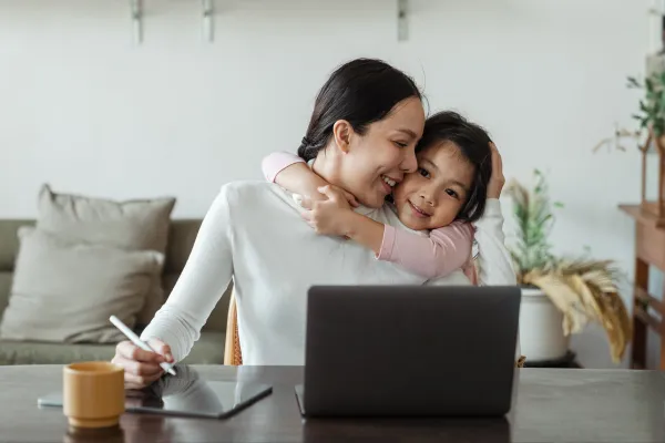 Mother working from home while her young daughter hugs her