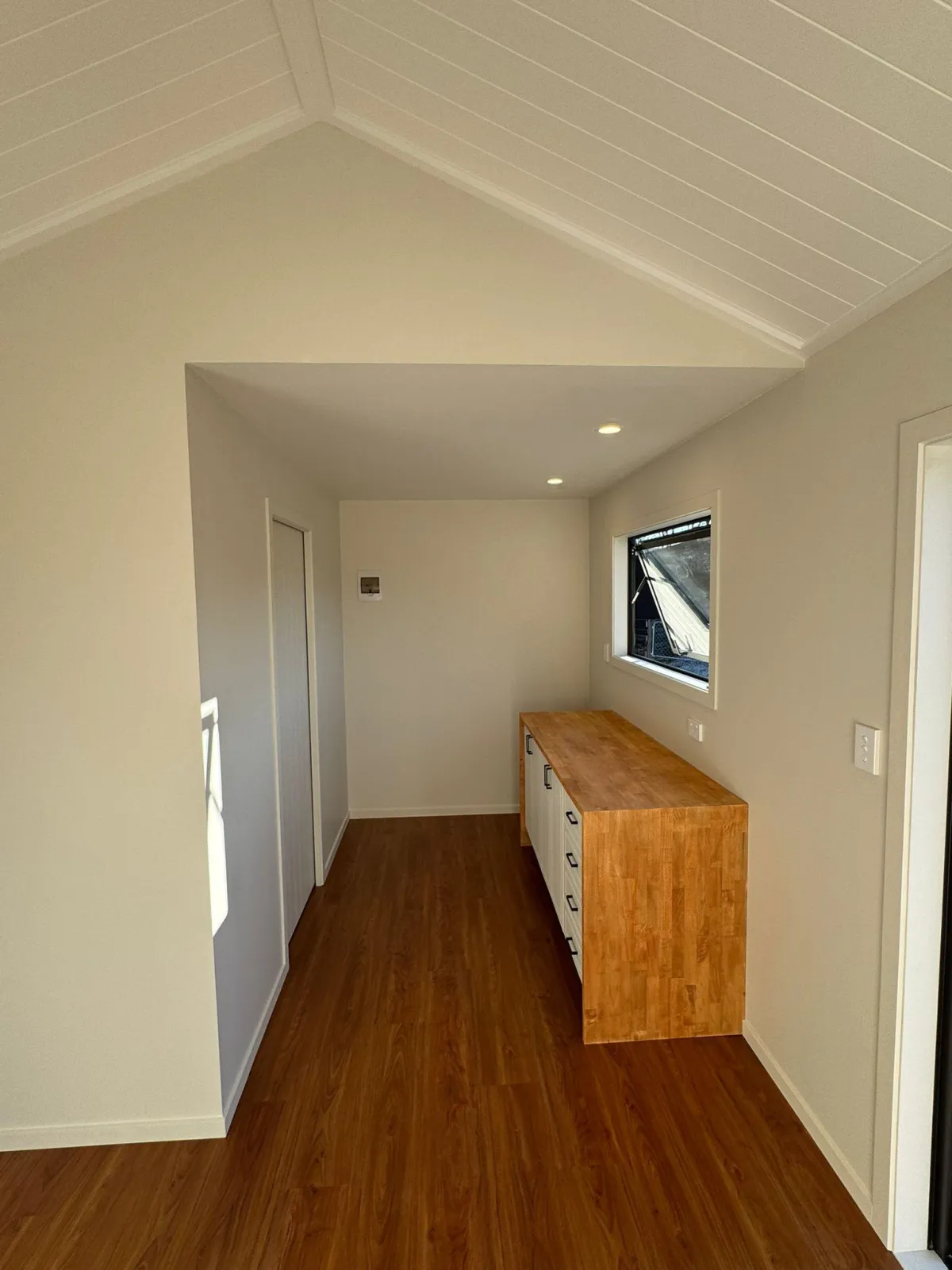 Vaulted ceiling interior of tiny home with timber kitchen cabinet and warm wood flooring.