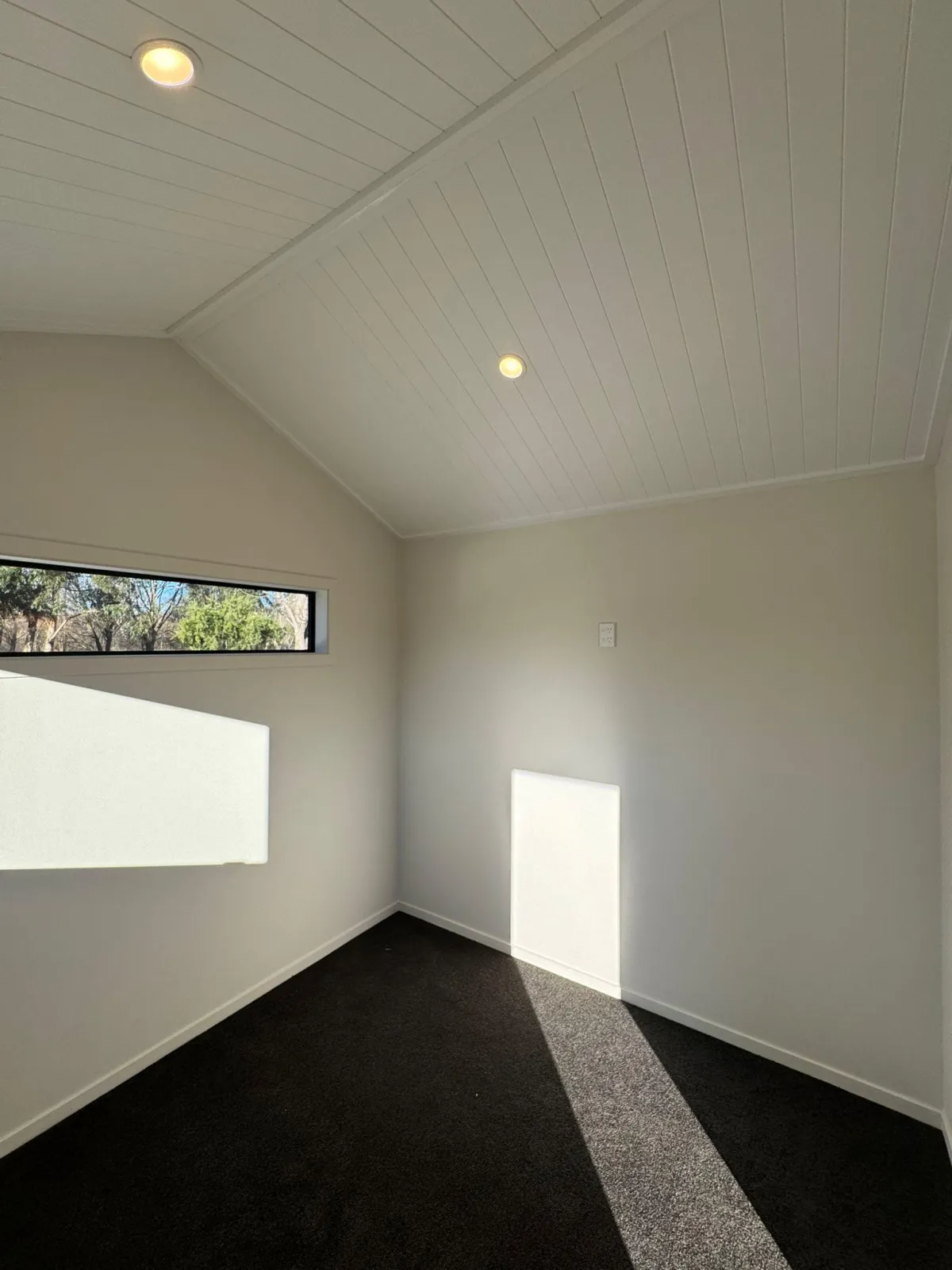 Carpeted sleepout room in tiny home with vaulted ceiling and natural light from horizontal windows.