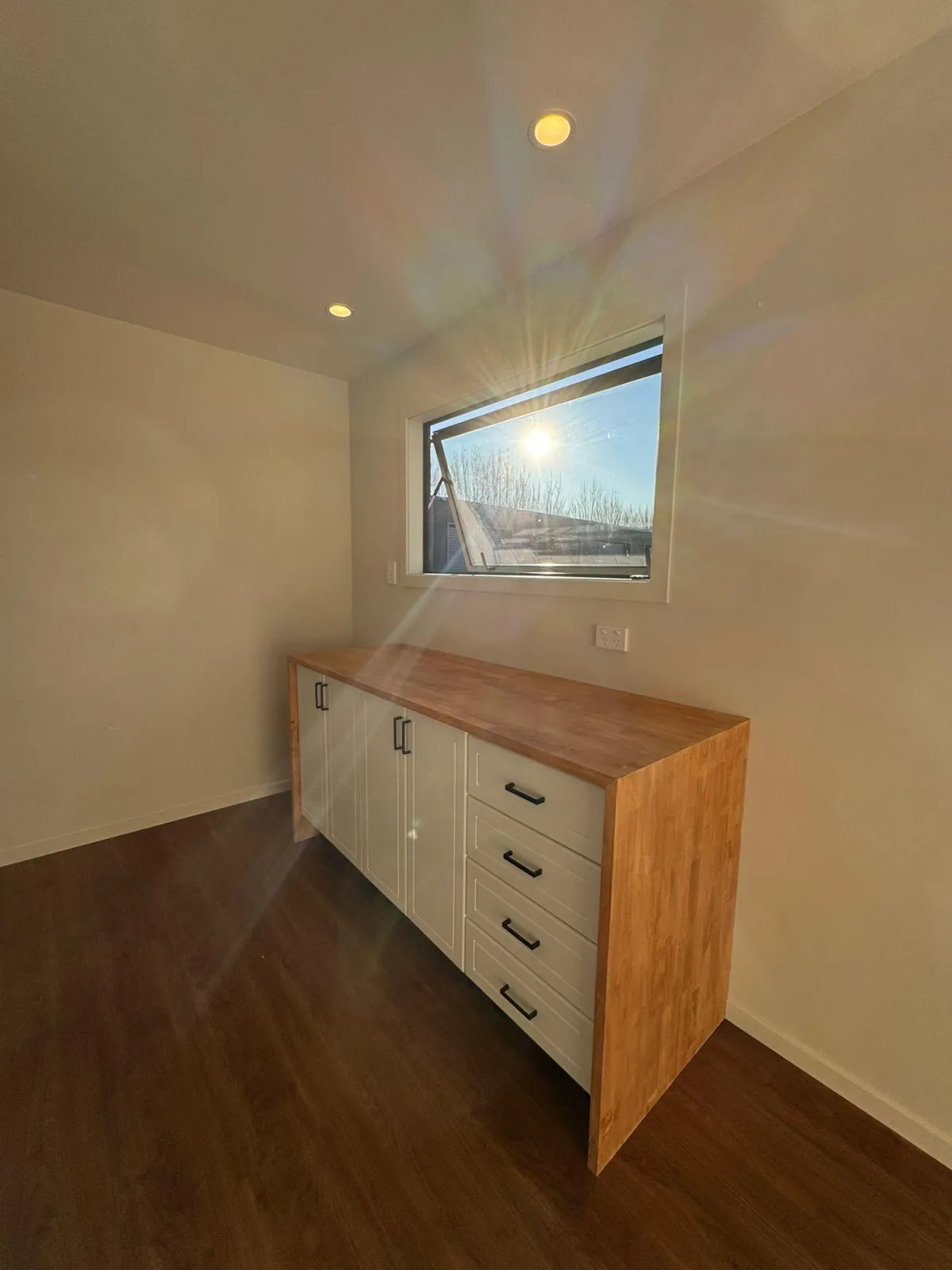 Timber kitchen cabinet with drawers and doors, sun shining through double-glazed window in tiny home.