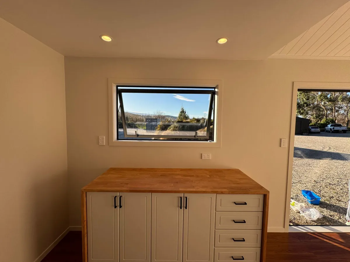 Wooden cabinet under large openable window inside compact cabin in Alexandra.