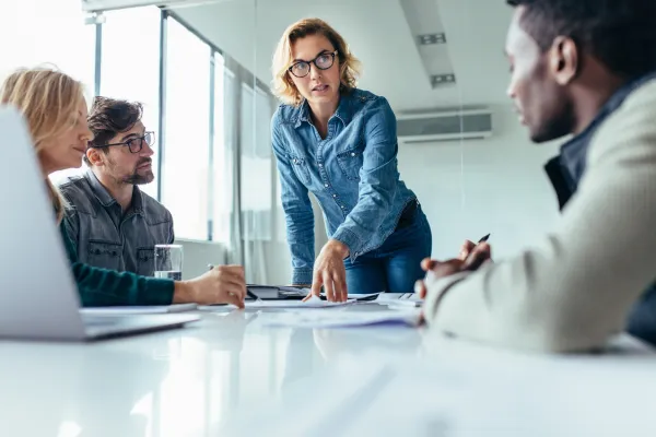 A woman in a denim blouse, in a corporate/office meeting, standing up and discussing a point to a male colleague