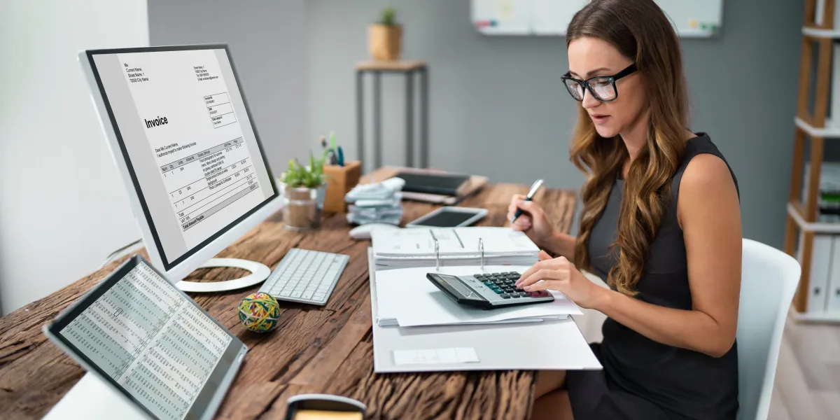 A lady sat at a desk with a calculator, an invoice on her screen and figures on her laptop