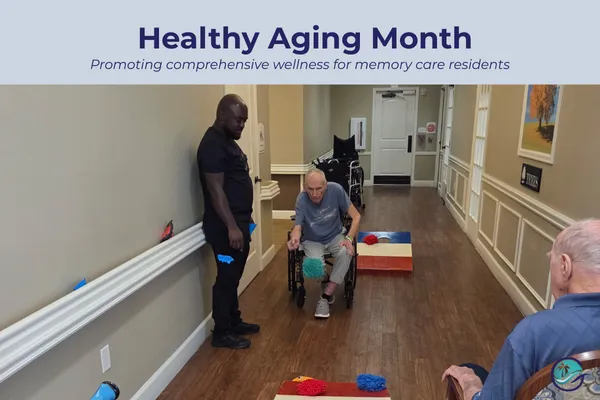 Residents at Gulf Coast Memory Care participating in an adaptive physical activity, with a staff member supporting a resident in a wheelchair playing a seated bean bag toss game in the community hallway, demonstrating inclusive and engaging wellness programming