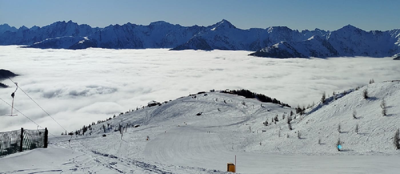 Blick vom Skigebiet Golzentipp über eine geschlossene Wolkendecke auf die verschneiten Gipfel der Karnischen Alpen unter blauem Himmel