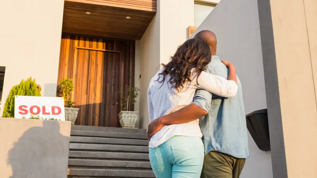 A couple stands arm in arm in front of their new home, looking at the entrance with a “SOLD” sign visible beside them. The image captures a moment of celebration and achievement after purchasing a house.
