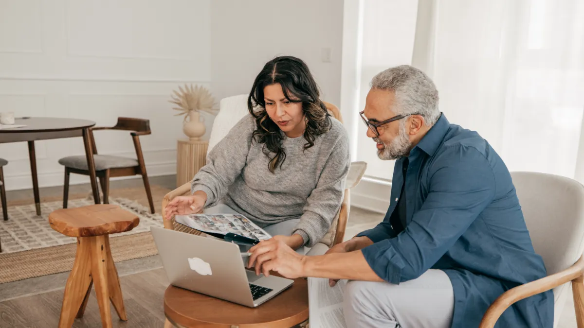 A mature couple sits together at a table in a cozy, modern living room, reviewing documents and looking at a laptop screen. They appear focused and engaged in a discussion, possibly planning a real estate decision or financial move.