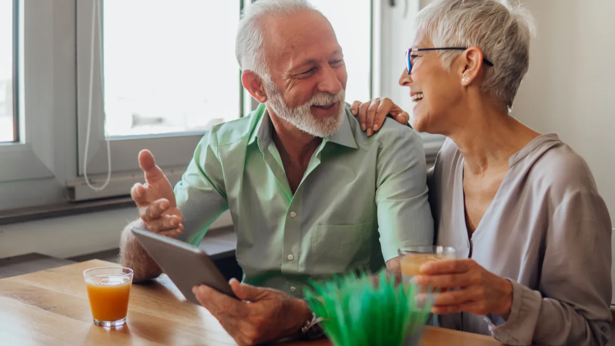 Smiling older couple sitting at a table together, enjoying a conversation. The man is holding a tablet while the woman rests her hand on his shoulder, both laughing warmly with drinks in front of them near a bright window.
