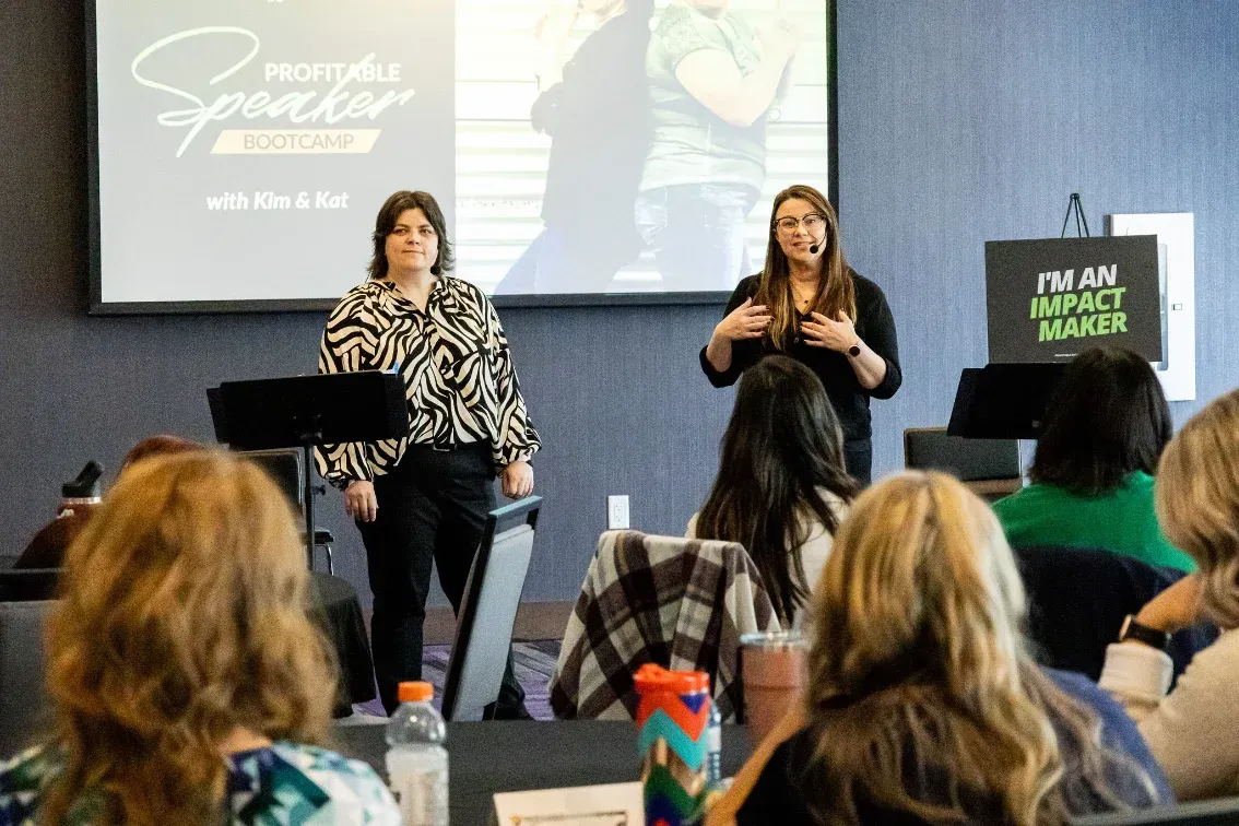 Kim and Kat presenting on stage at the Profitable Speaker Bootcamp with attendees seated and event signage visible.