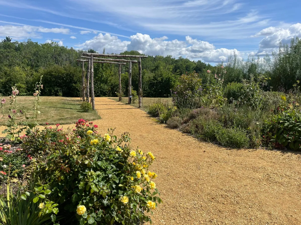 Scenic garden pathway with blooming yellow and pink flowers, wooden archway, and lush greenery under a bright blue sky with clouds