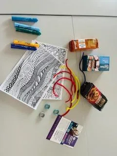 Flat lay of neurodiversity workshop materials on a grey table, including colouring sheets with abstract patterns, colourful felt-tip pens, fidget rings, pipe cleaners, snacks, and informational leaflets.