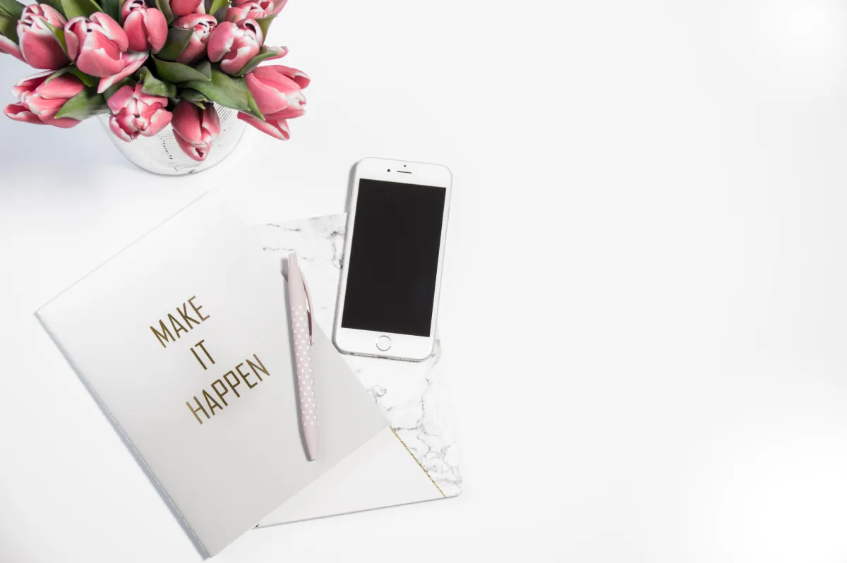 Whitdesk with a phone and a book with "Make it Happen" on the cover and some pink tulips
