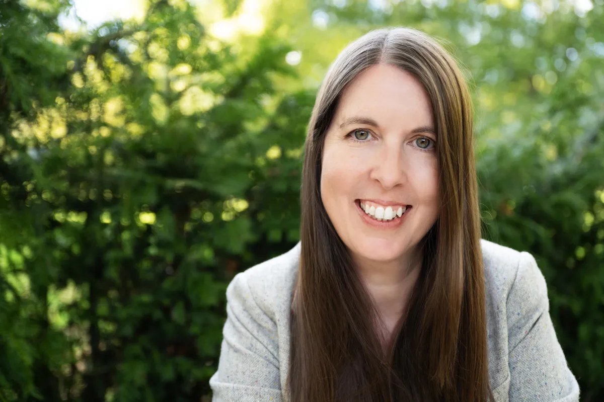 Photo of Sarah smiling at the camera with a grey blazer and surrounded by green trees