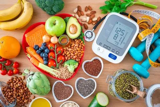 Heart care concept. Overhead view of a group of food that contains healthy nutrients for heart care, a blood pressure monitor, dummbells, tape measure and sthetoscope shot on wooden table. The composition includes extra virgin olive oil, legumes, avocado, berries, nuts and seeds, salmon, broccoli, carrot, soy beans, brown lentils, eggplent, spinach, oats, artichoke, apple, tomato, garlic among others.