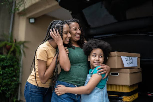 Three people embracing beside an open car trunk filled with boxes labeled “DORM ROOM,” standing outside a residential building with greenery in the background—capturing a heartfelt moment of support and transition, symbolizing the emotional strength and connection behind Self-Love Mantras.
