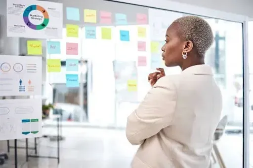 A person stands in front of a glass wall filled with colorful sticky notes and strategic charts, deeply engaged in planning and analysis. The vibrant workspace reflects themes of clarity, intention, and growth—visually echoing Self-Empowerment Mantras: 7 Soulful Ways to Step Into Your Power through focused action, vision mapping, and confident decision-making.