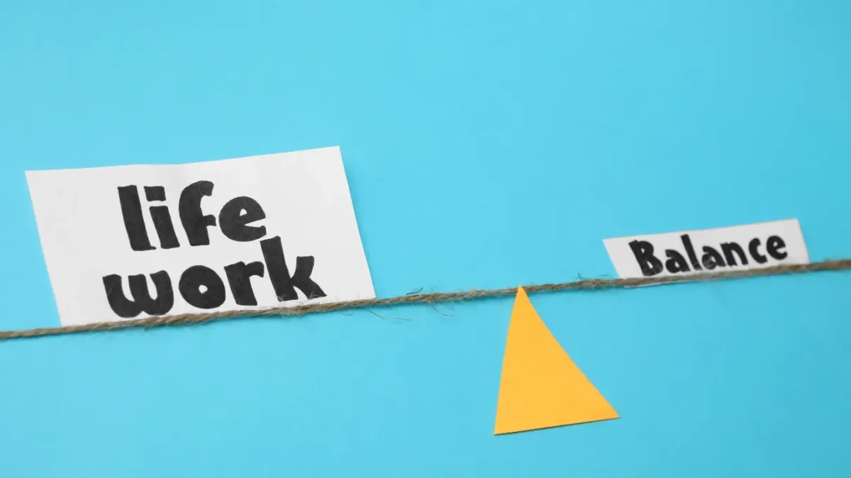 Conceptual image of work-life balance featuring a string stretched across a yellow triangle fulcrum, with papers labeled “life,” “work,” and “balance” on a blue background—symbolizing Career Growth Without Burnout: Balancing Ambition and Wellbeing through equilibrium between personal and professional priorities.