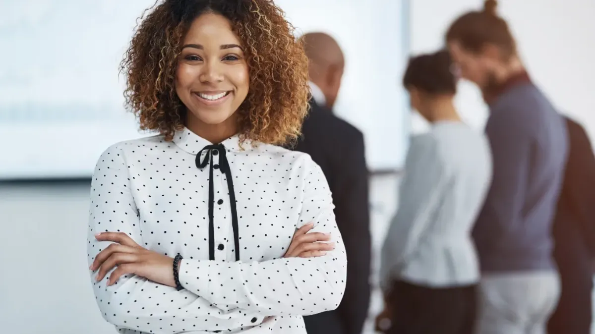 Confident professional in a white polka-dot blouse standing in a bright office setting, symbolizing empowered leadership and workplace presence. Balanced Leadership: Work, Wellbeing, and Purpose.