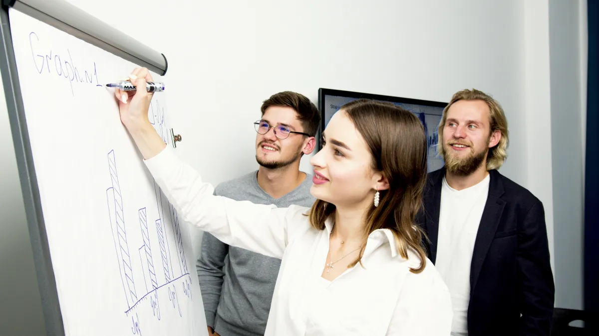 A young female insurance agent drawing a graph on a flip chart, with two male colleagues, one with glasses and one with a beard, standing behind her, discussing the challenges and growth opportunities of offering Medicare products.