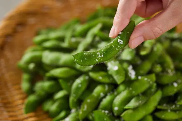 a hand holding an edamame bean above a bowl of edamame beans