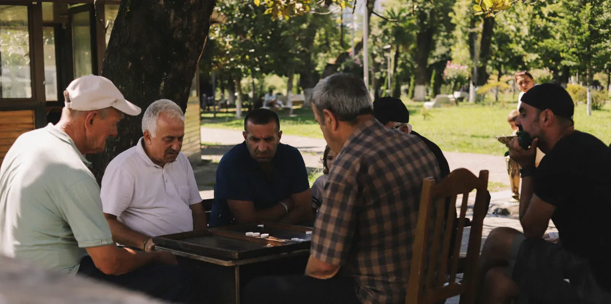 Group of men sitting outdoors at a park table playing a board game.