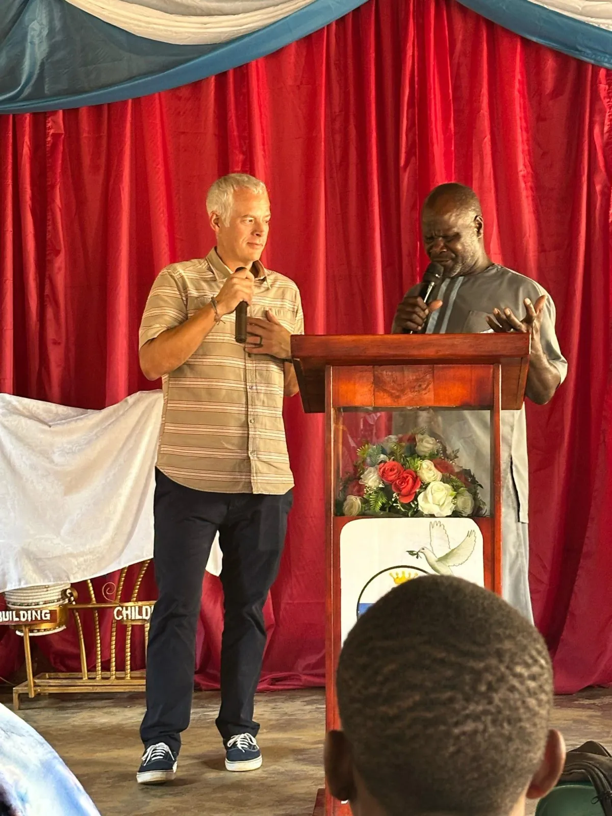 Two men, one Caucasian in a striped shirt and the other Black in a gray traditional outfit, stand at a wooden podium speaking into microphones.