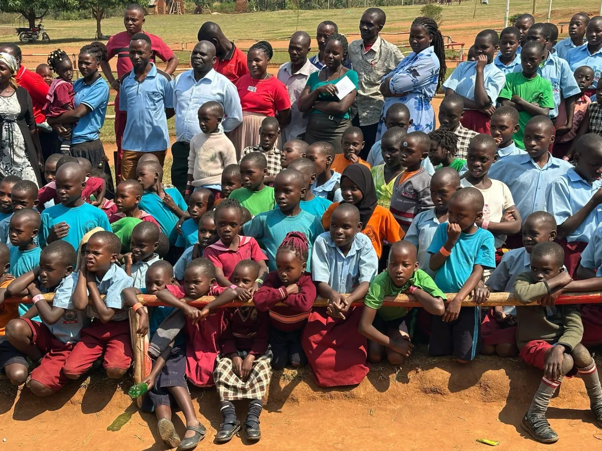A large group of children, mostly boys, and some adults are gathered outdoors on a sunny day.